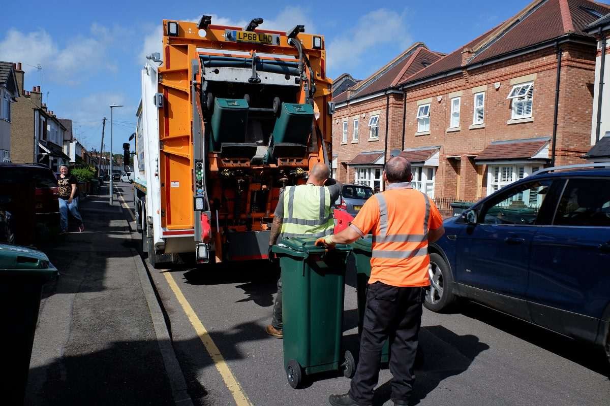 Bin lorry on urban street in England with waste operatives nearby, pushing wheelie bins
