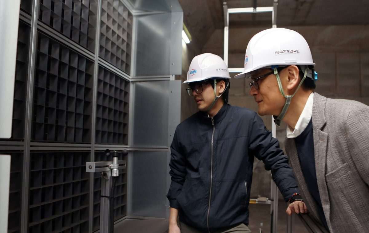 Two men in hard hats crouch to inspect T-shaped metallic assembly, in an industrial environment