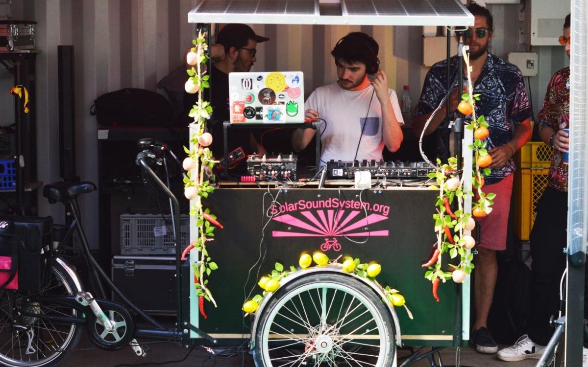 A photo of a DJ with headphones and a laptop standing inside a small booth with a bike wheel on its front and solar panels on top of it