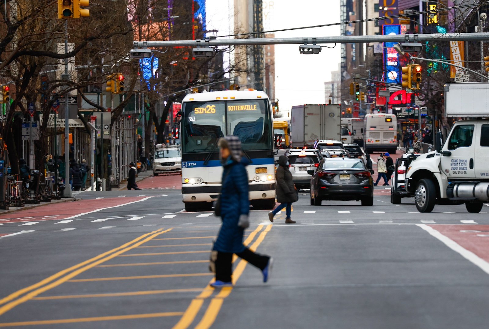 A person wearing black jaywalks across a city street with a municipal bus and several cars and trucks behind them on a cloudy day