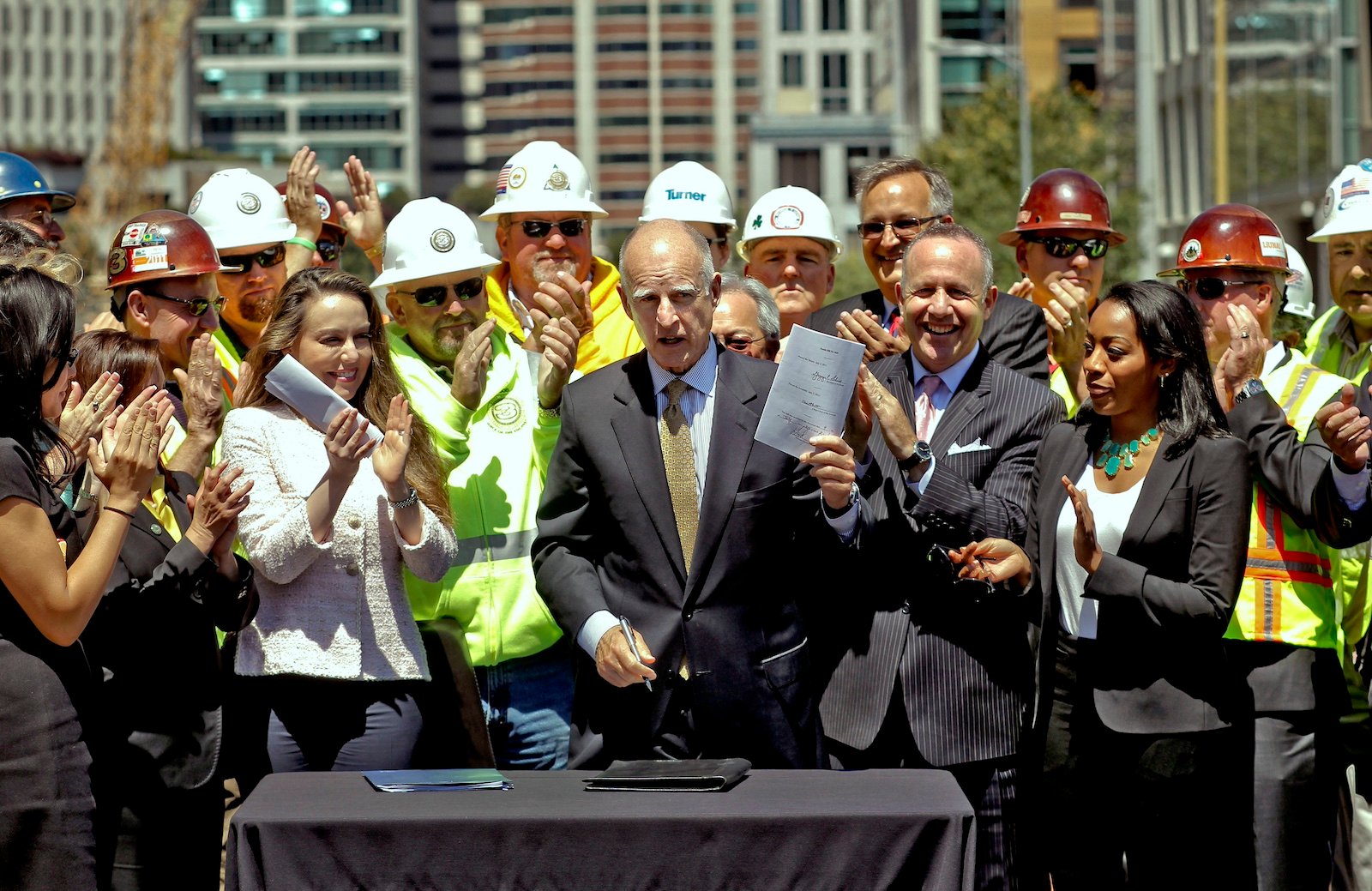 A bald man in a suit stands behind a desk, outdoors, holding a packet of papers and a crowd of people wearing hard hats around him applaud