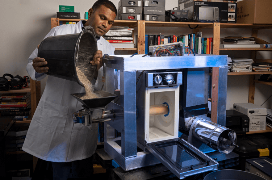 Scientist in a building interior pouring powder-like material from a bucket into a tray attached to a metallic assembly with cylindrical attachments, resembling a futuristic microwave