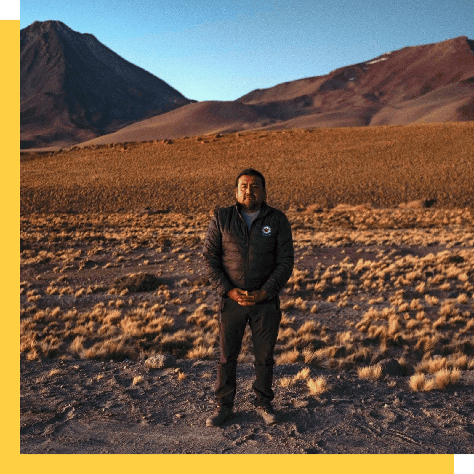 A cropped photo of a man standing in the middle of a desert with blue sky behind him. The crop is a square and highlighted by a golden design edge