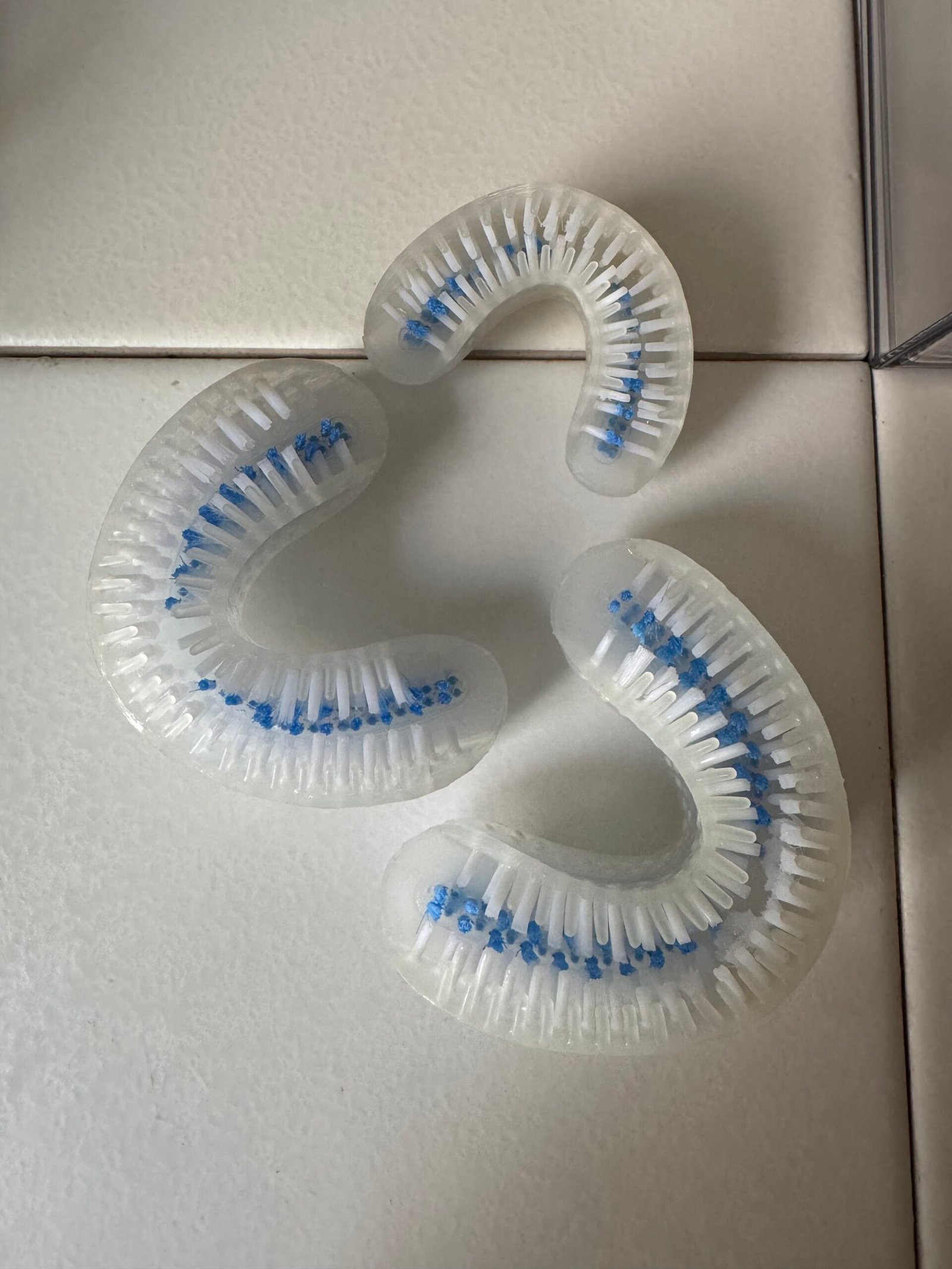Three curved, clear plastic teeth-whitening mouthpieces with blue bristles are arranged on a white tiled surface.