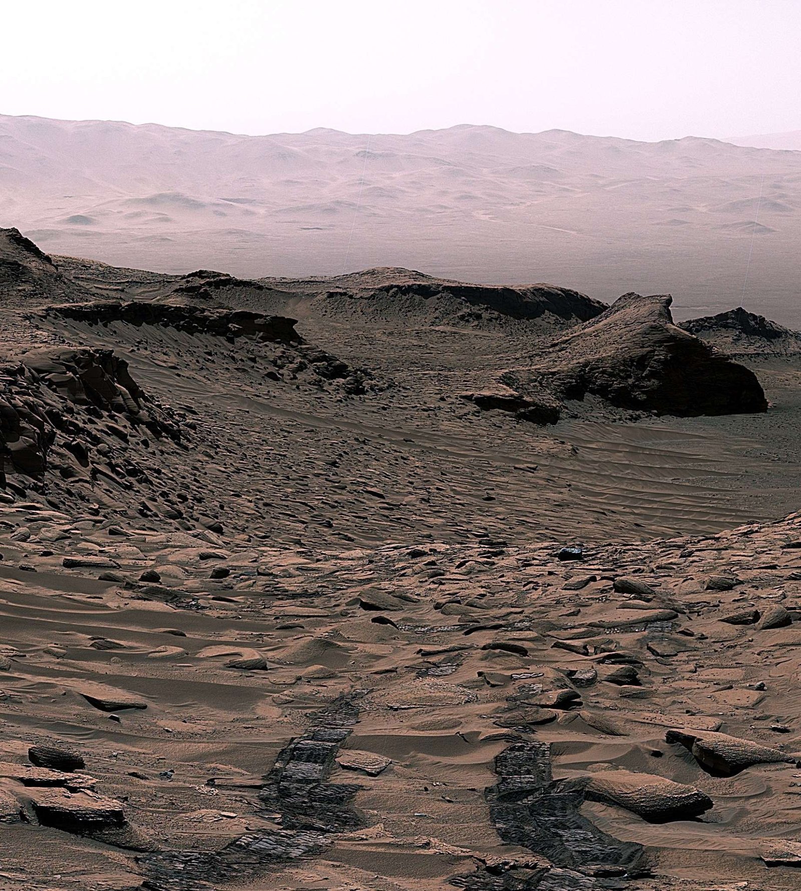 An image of Mars shows rocks and small sand dunes, with a larger mountain range in the background. Two wheel tracks in the foreground show Curiosity rover's path before it captured the image.