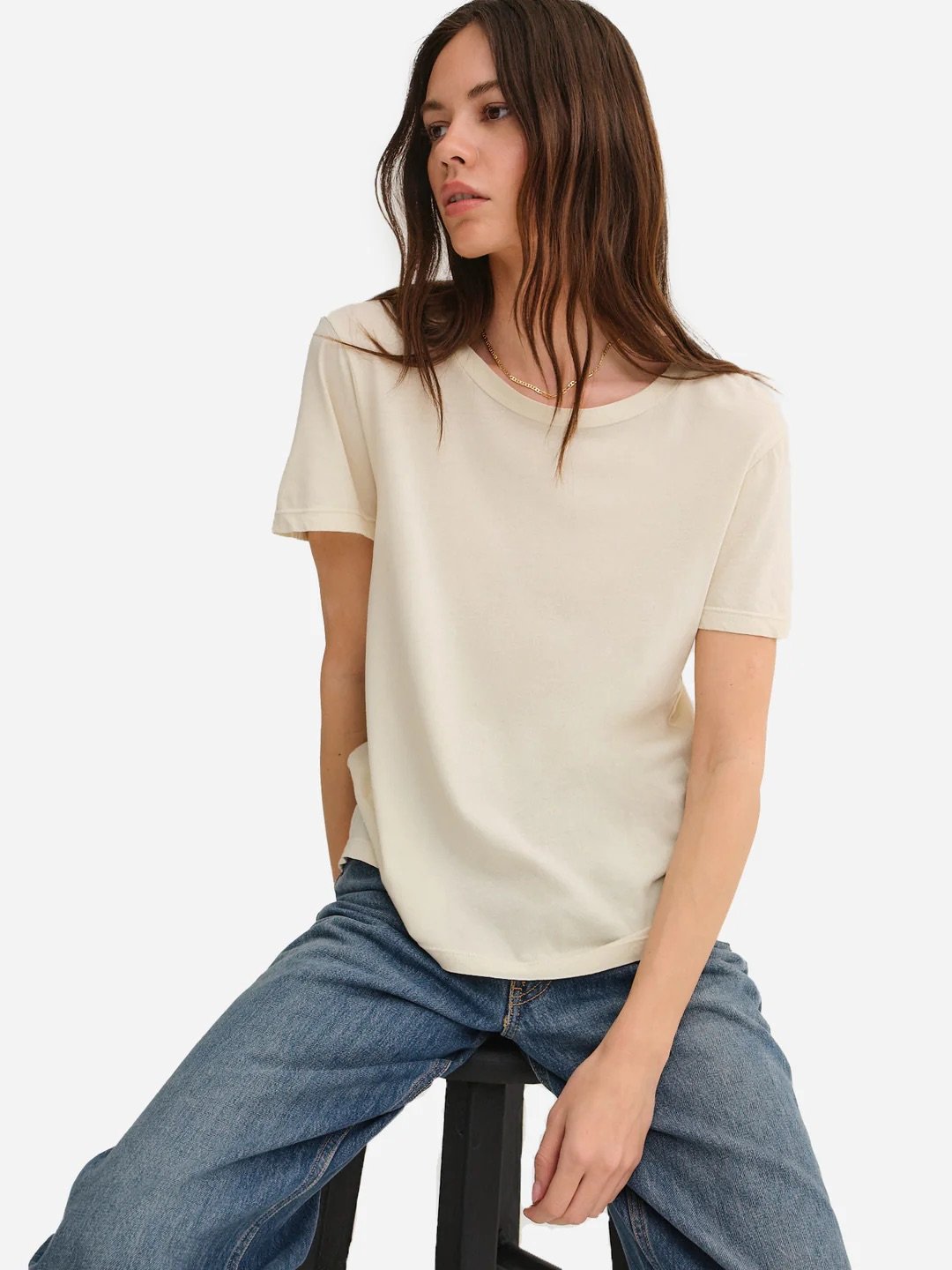 Person with long hair, wearing a cream-colored t-shirt and blue jeans, sitting on a stool against a white background.