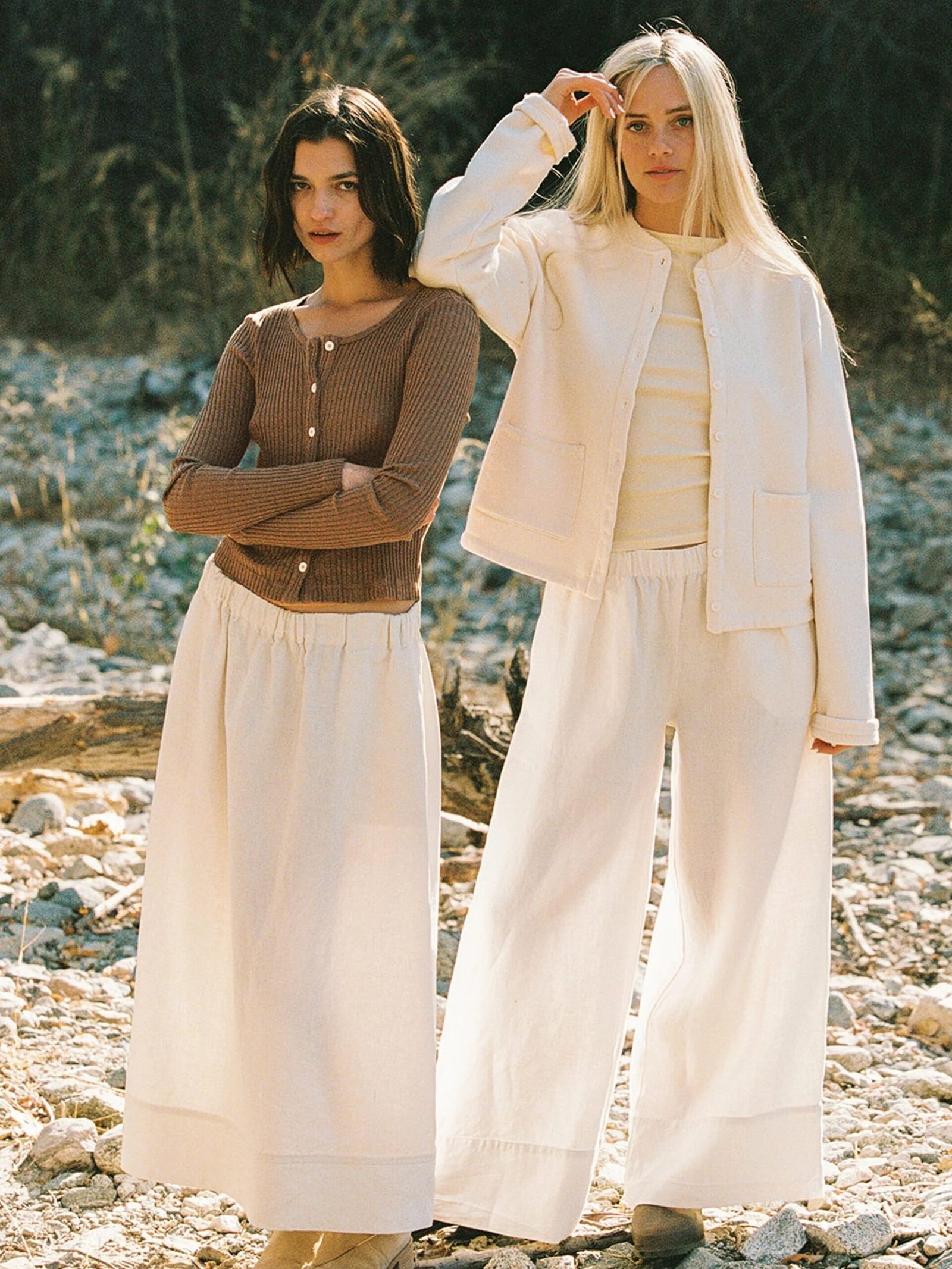 Two women stand in a rocky outdoor setting. One wears a brown top and cream pants, while the other is in a cream outfit with a jacket. Trees are visible in the background.