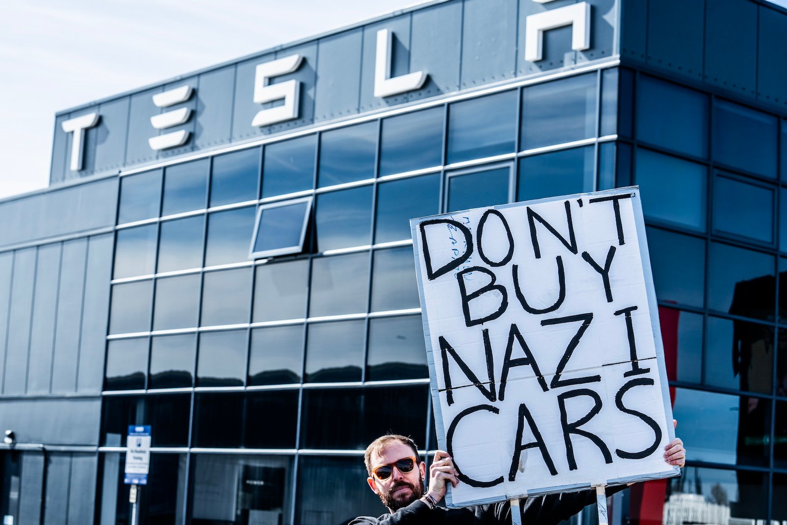 A person wearing sunglasses holds a sign that says 'Don't Buy Nazi Cars' in front of a glass building with the Tesla logo on it