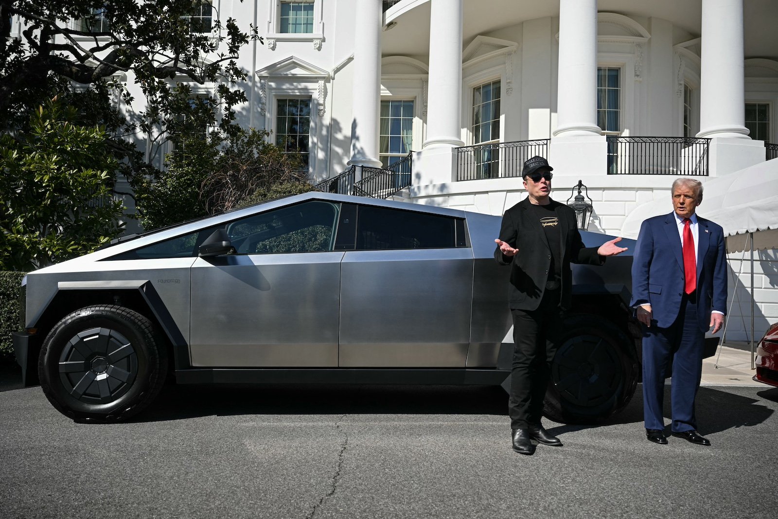 Elon Musk, in a black baseball cap and sunglasses, stands next to Donald Trump, in a blue suit with a red tie, in front of a Tesla Cybertruck in front of a grand white-columned building