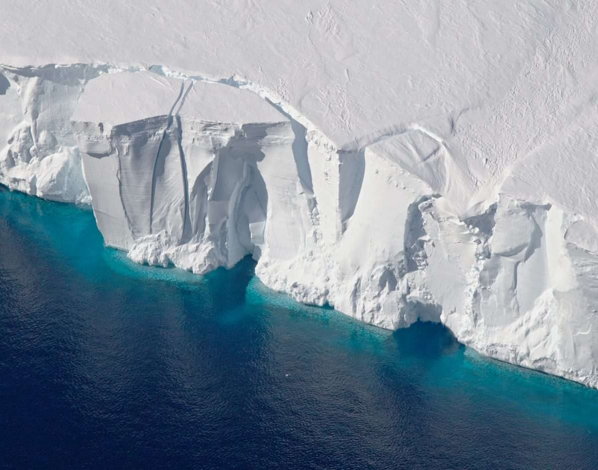 Aerial view of the crumbling edge of a large ice mass, or ice shelf, as it meets the sea, which is coloured turquoise near the ice but blackens as it moves further away from it
