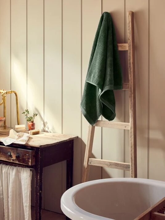 A bathroom with a wooden ladder holding organic towels, a rustic sink with a gold faucet, a round white bathtub, and a lit candle on the tub edge.