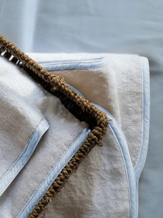 A close-up of folded organic towels with blue trim inside a woven basket, placed on a light blue surface.