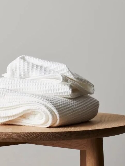 A set of two white organic towels with a waffle texture is folded on a wooden table next to a beige ceramic bottle and a small brush.