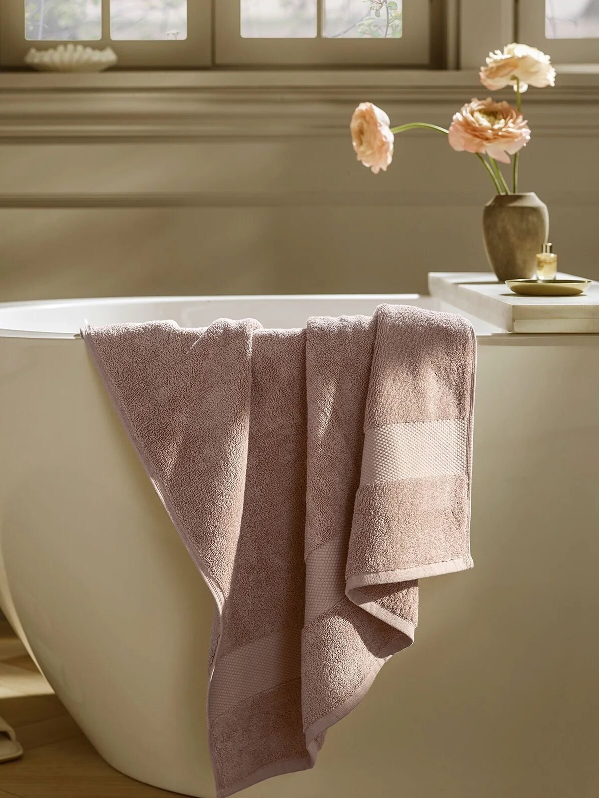An organic towel hangs over the edge of a white freestanding bathtub, with slippers on the floor and a vase with flowers on the ledge nearby.