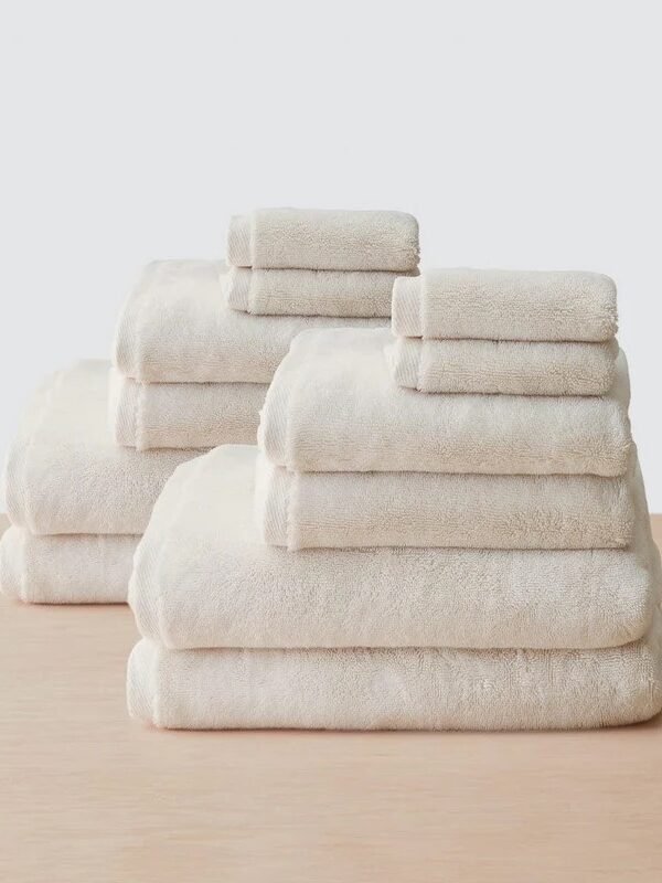 Two stacks of neatly folded, cream-colored organic towels in various sizes are arranged on a light wooden surface against a plain background.
