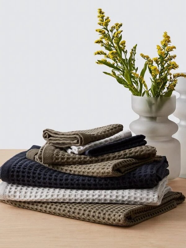 Stack of folded organic, waffle-textured towels in neutral colors on a wooden surface, with two white vases holding yellow flowers in the background.