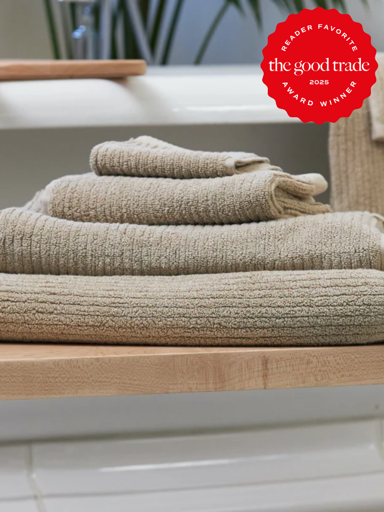 A stack of beige organic towels folded neatly on a wooden shelf, with a red "The Good Trade 2025 Award Winner" badge in the upper right corner.