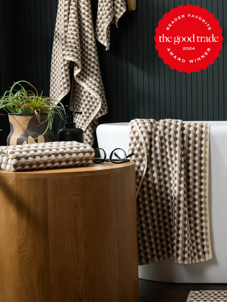 A modern bathroom features organic checkered brown and white towels, a wooden table with a plant and glasses, and a white bathtub. A red "The Good Trade 2024 Award Winner" badge is displayed.