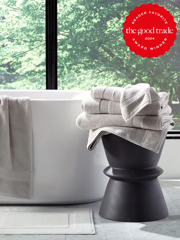 A modern bathroom with a white bathtub, organic gray towels stacked on a black stool, a gray bath mat, and a large window overlooking greenery. "Reader Favorite" award badge in upper right corner.