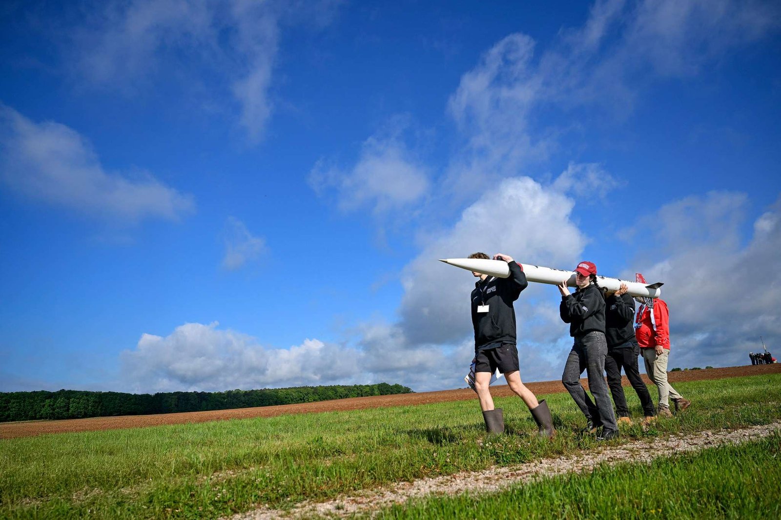Four people carry a small, thin white rocket across a grassy field. The photo is taken from a lower angle, so the blue sky stretches out over most of the background.