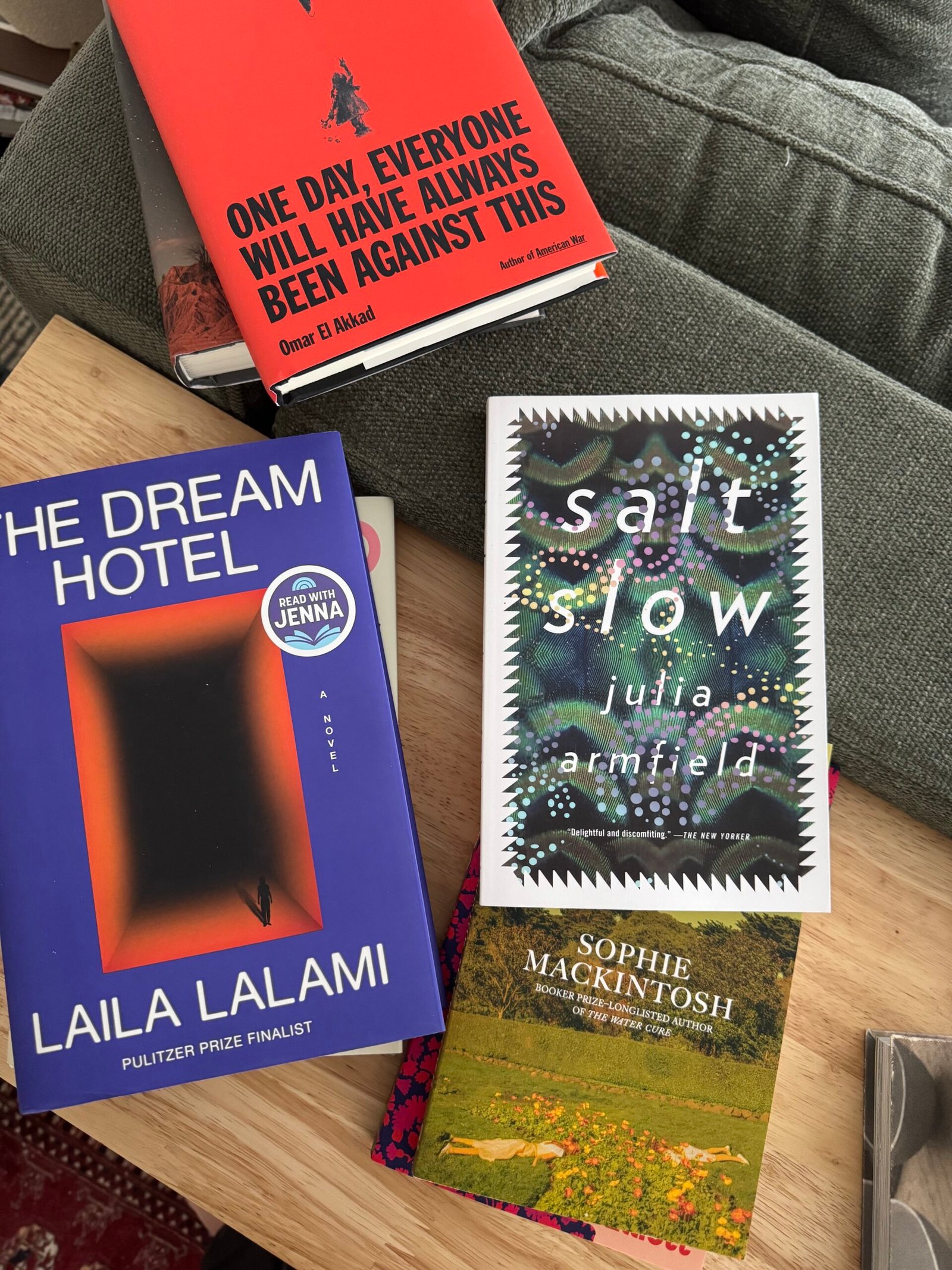 Four books—"The Dream Hotel," "Salt Slow," "One Day Everyone Will Have Always Been Against This," and "Cursed Bread"—are arranged on a wooden table near a green sofa cushion.