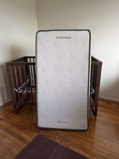 A white Avocado brand infant mattress stands upright in front of a wooden crib in a room with wood flooring.