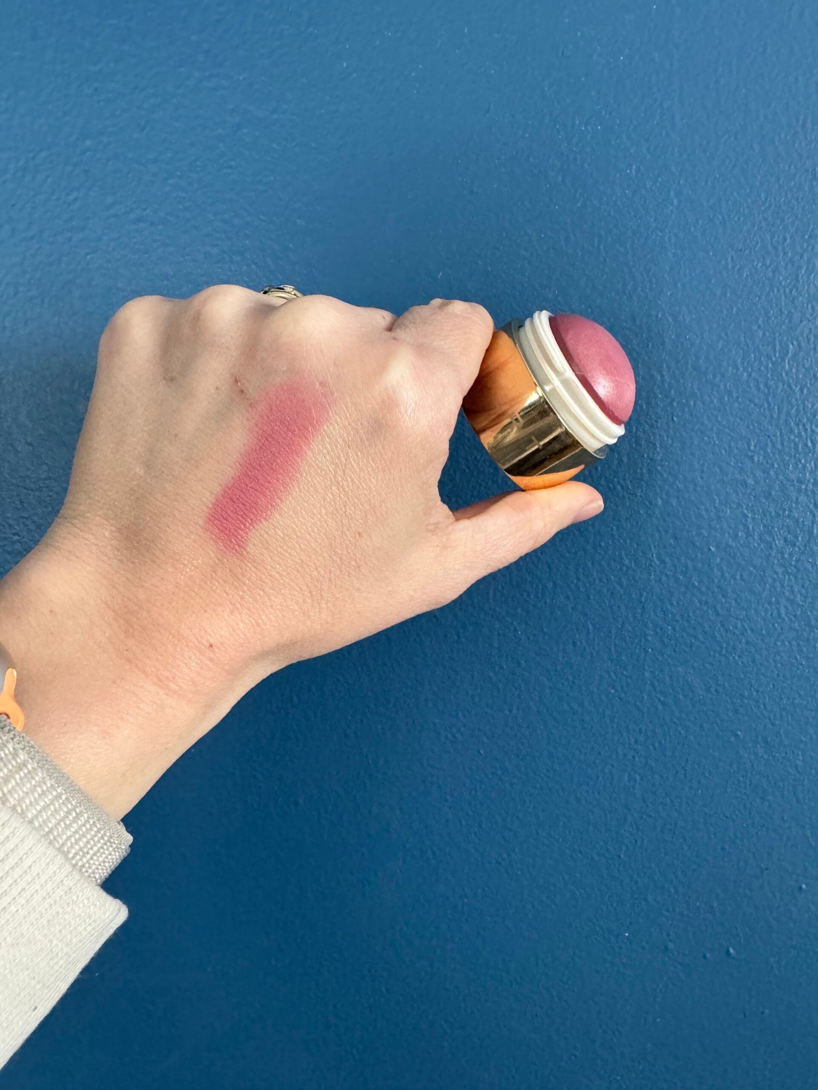 A hand holding a cosmetic product against a blue background with a swatch of pink makeup on the skin.
