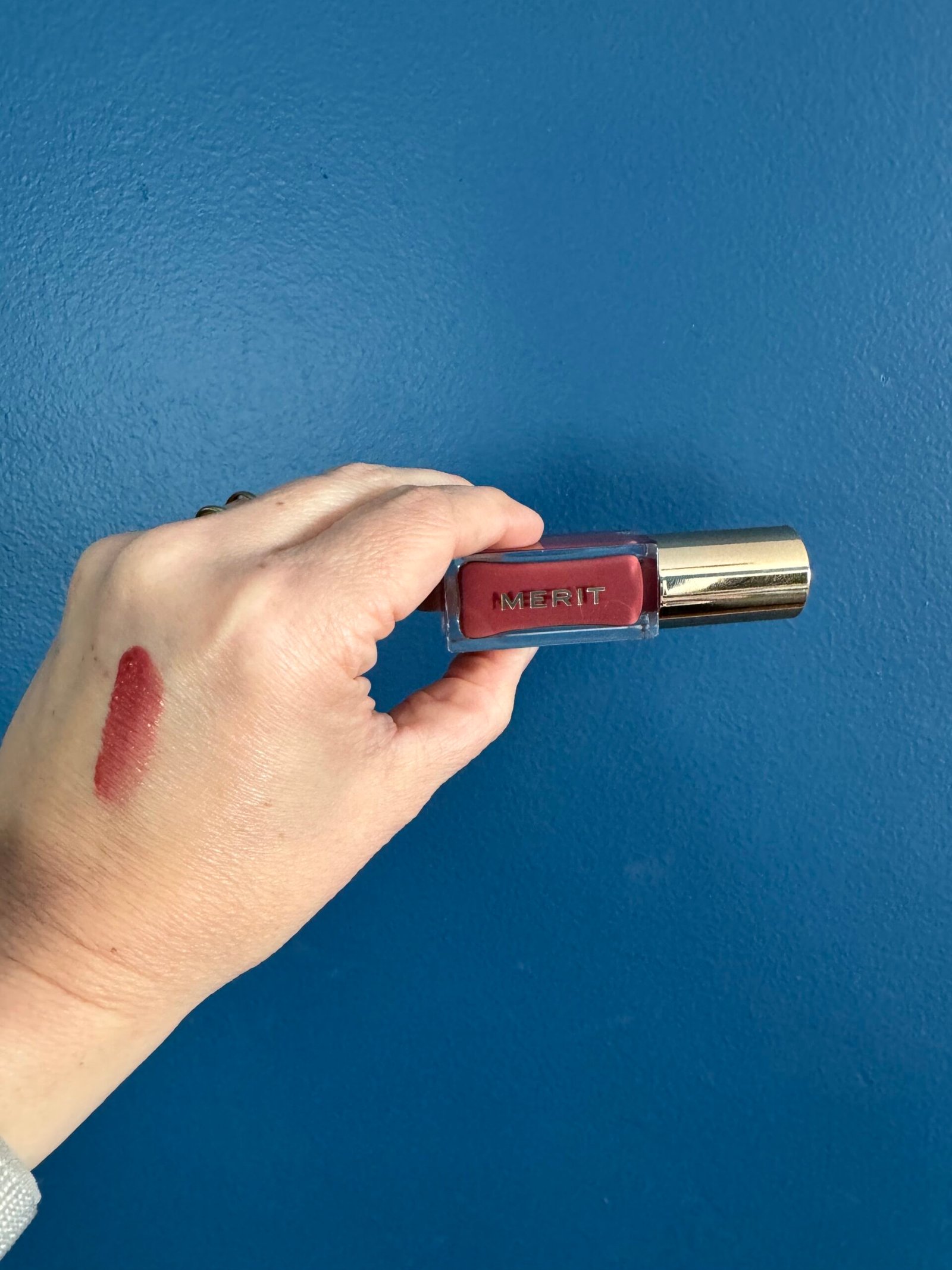 A hand holding a tube of merit lipstick against a blue background, with a swatch of the lipstick color on the person's wrist.