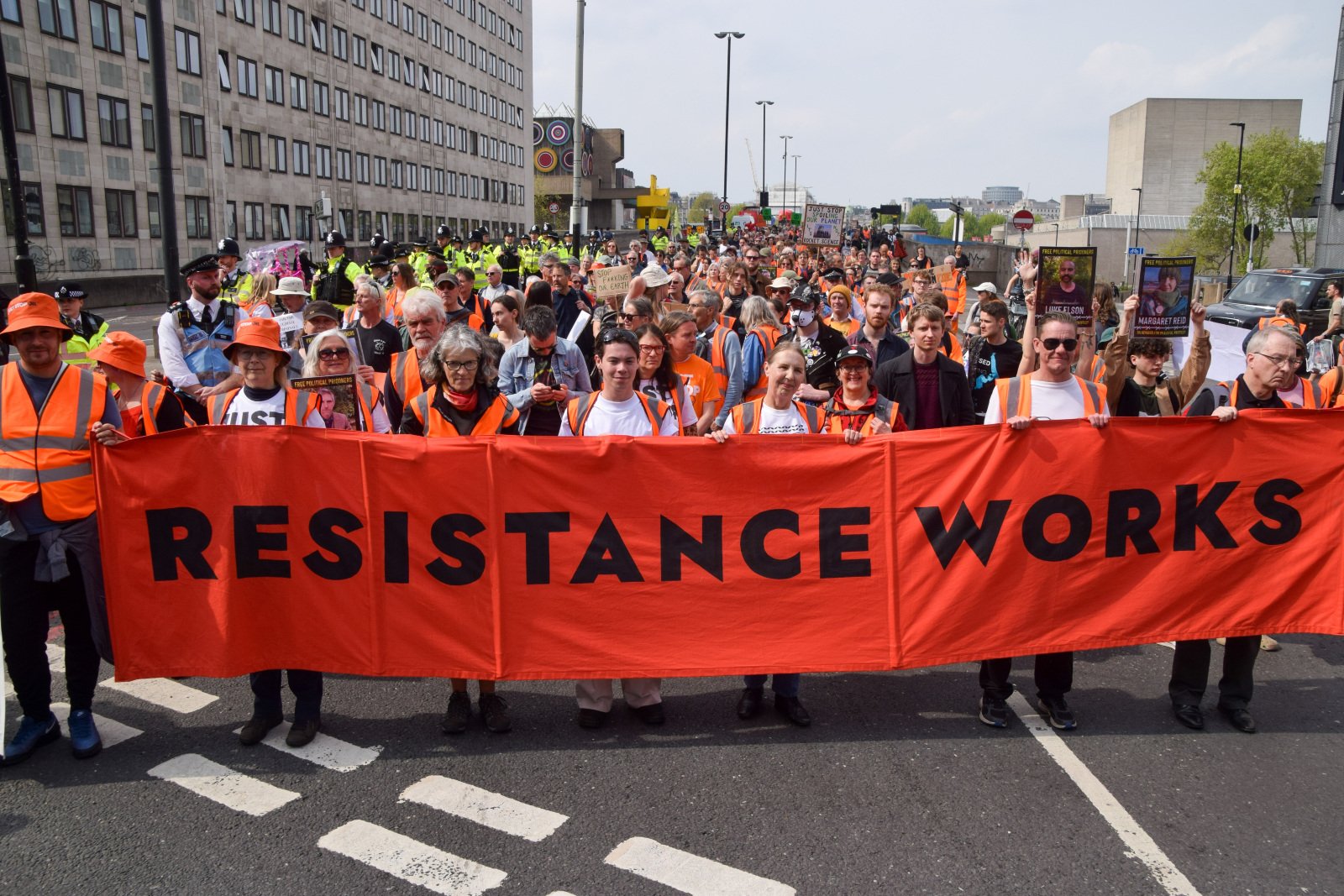 A crowd of people march in a road, holding a sign saying "resistance works"