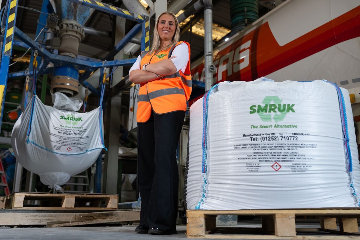 A woman in luminous safety vest stands next to a palletised bag on which is printed the words 'SMRUK'