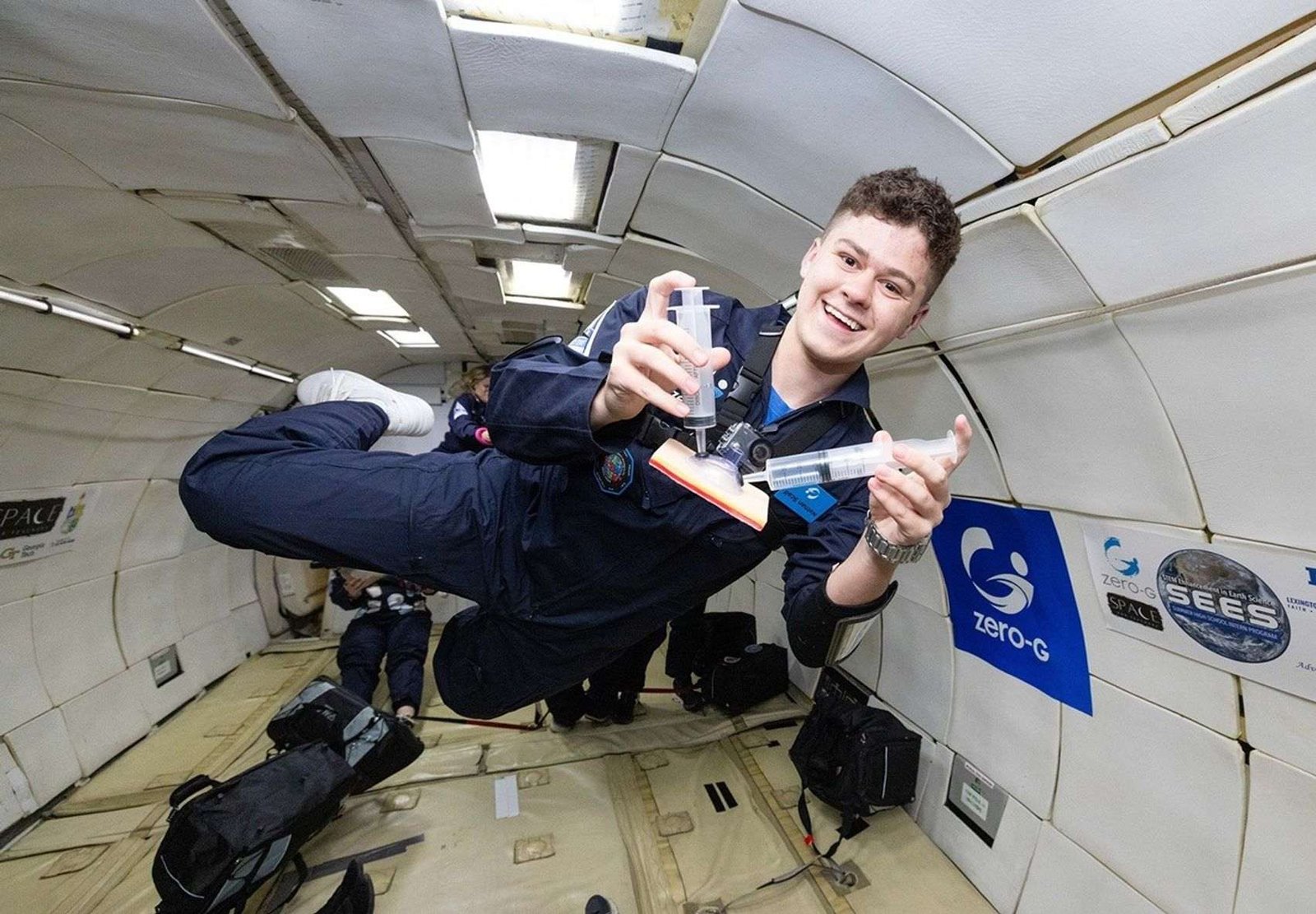 A young man in a navy blue flight suit floats inside a microgravity simulation aircraft, smiling as he demonstrates a Wound Irrigation System using two syringes and a small device. The padded interior of the aircraft is visible behind him, along with logos for Zero-G and NASA SEES on the wall. He appears weightless, mid-air, during a parabolic flight aboard the ZERO-G G-FORCE ONE®.