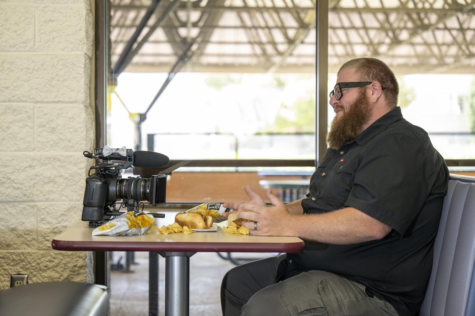 A man with red hair and a beard, wearing black-rimmed glasses and a black short sleeved shirt, gestures as if in conversation. He is seated on the right side of a cafeteria booth, pretending to talk to his camera, which sits across from him on the table. A hot dog in a bun, two bags of chips, and scattered chips are on the table between them. The photo is playfully staged to look like a lunch meeting between the man and his camera.