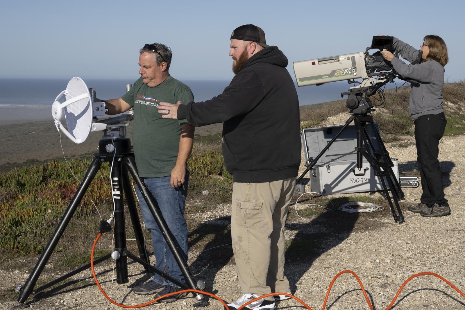 Two men stand behind camera equipment, preparing to film a scene outside the left side of the frame. One, in a green short-sleeved shirt and jeans with sunglasses on his head, is in the foreground; the other stands slightly behind and to the right, wearing a backward brown-and-black baseball cap, a black hoodie, and khaki pants. An orange cord is plugged into the equipment on a tripod and spirals along the ground, exiting the frame on the right. Behind them, one the right side of the frame, a woman in a gray long-sleeved shirt and black pants adjusts a camera on a tripod. The team is on a dirt path bordered by beach plants, with sand, ocean, the horizon, and blue sky in the distance.