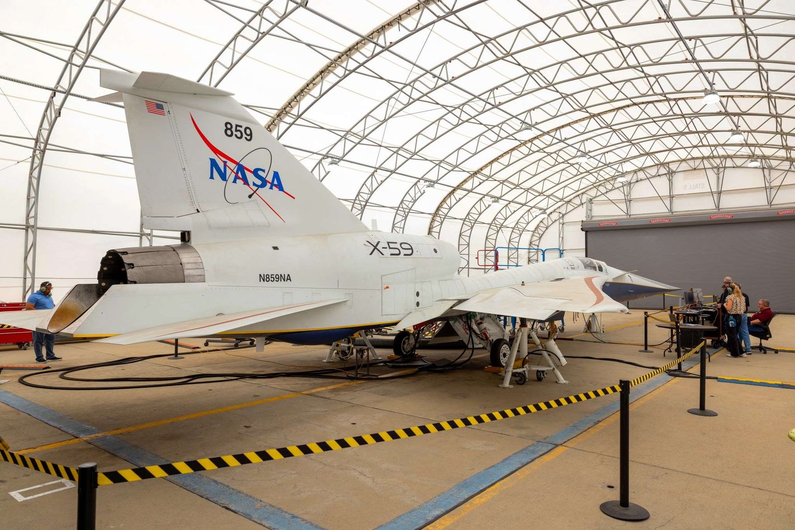 A white NASA jet airplane sits inside a fabric-covered-roof hangar with several people working at a nearby table with computers.