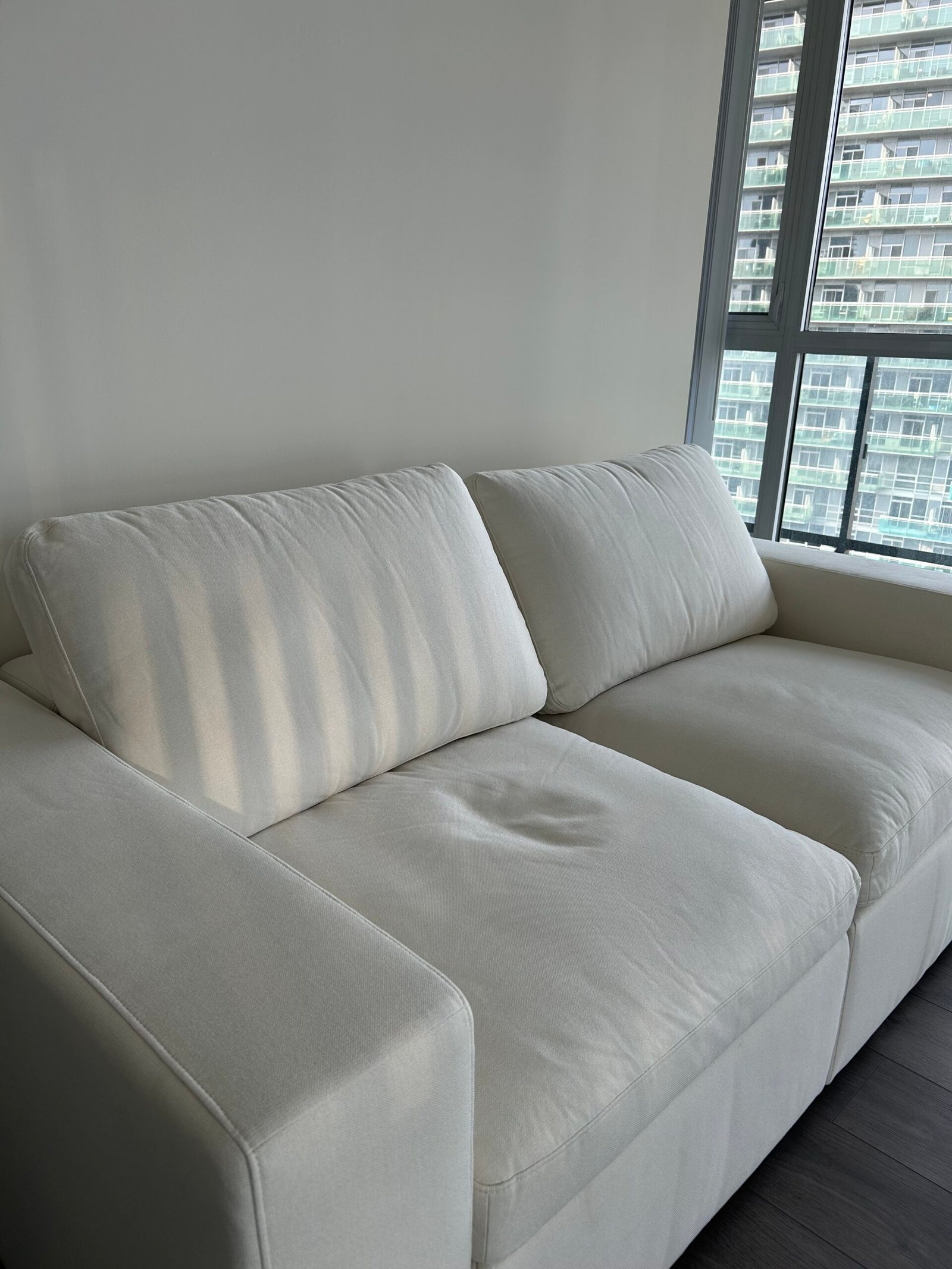 A white, modern two-seat sofa sits near a large window with vertical blinds, in a bright room with dark wooden floors.