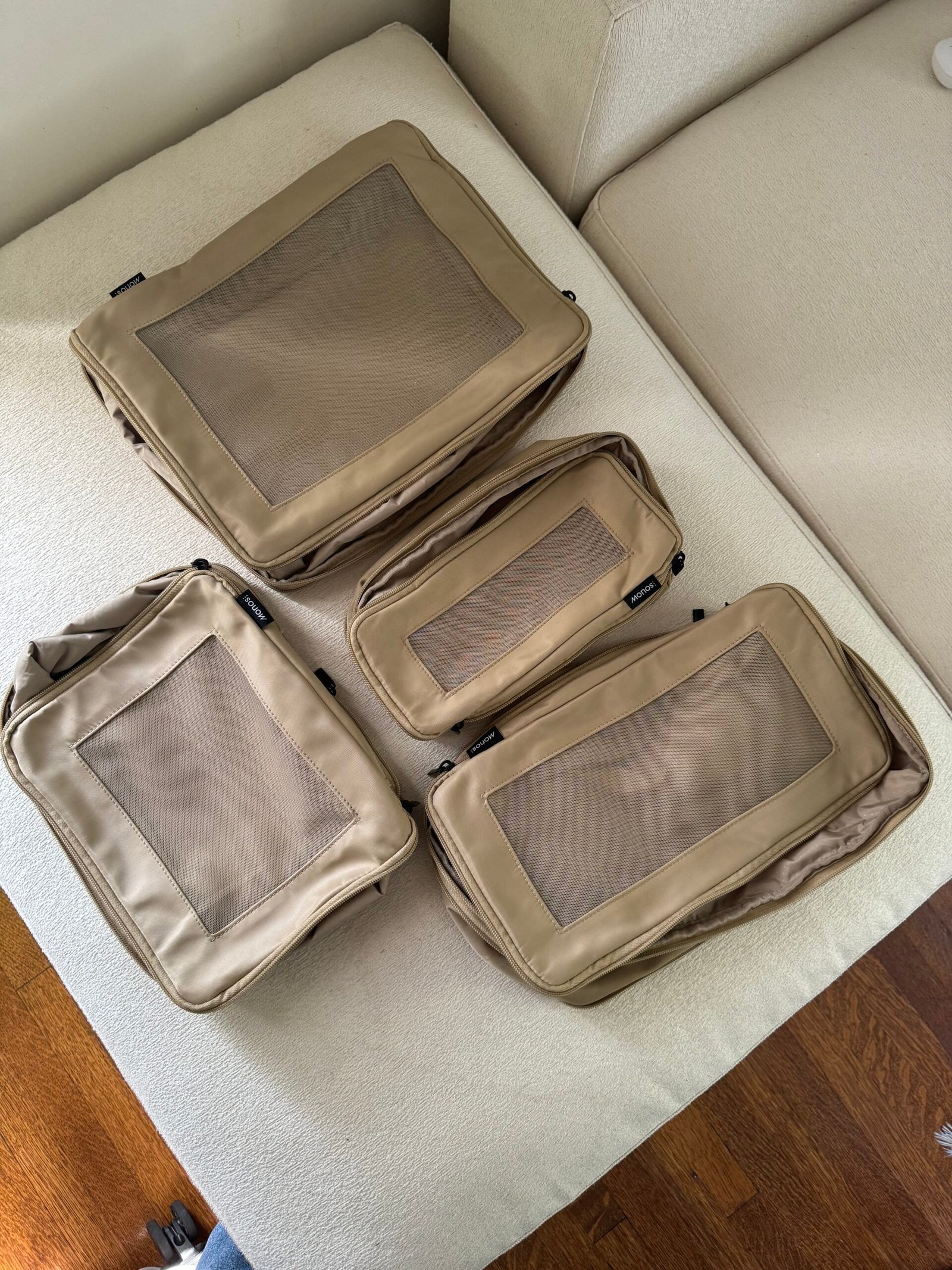 Four tan packing cubes of varying sizes are neatly arranged on a cream-colored sofa, with part of a wooden floor visible in the corner.