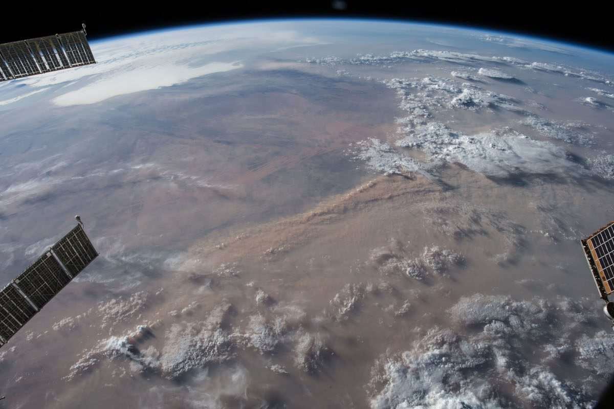 A segment of the Earth's surface as seen from orbit, incorporating the curvature of the planet as it meets the horizon of space, with details like clouds and brown or ochre desert-like surface faintly visible