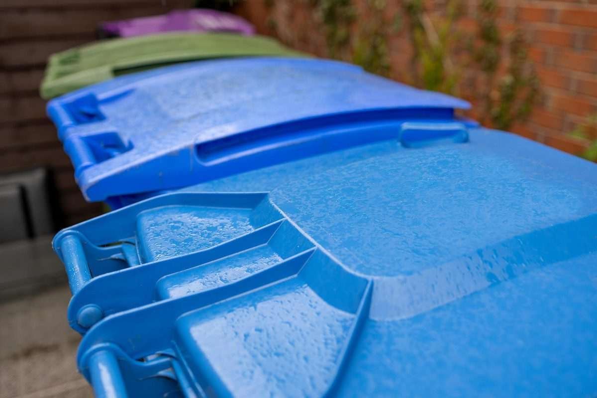 Coloured wheelie bins arranged side by side, seen from slightly above and to the side, so that each successive bin appears further away from the camera