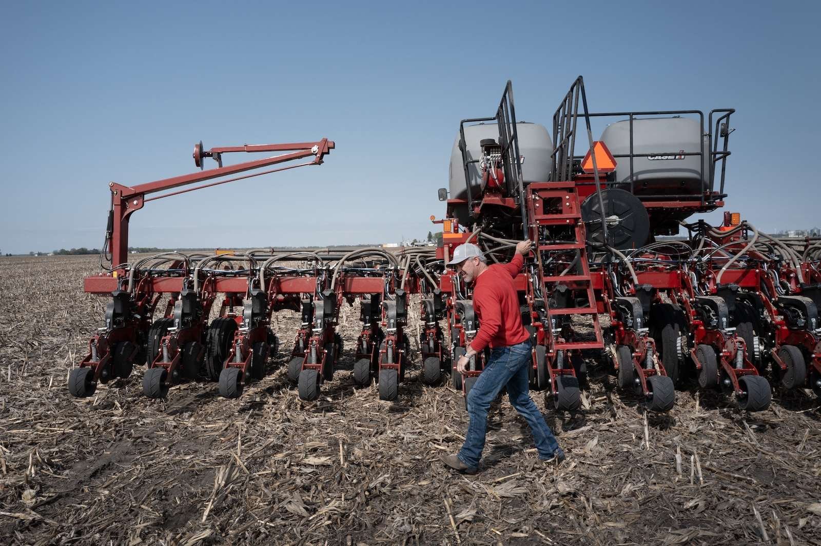 a farmer dressed in a red long-sleeve shirt and jeans walks in front of a red tractor tilling soil with blue sky overhead