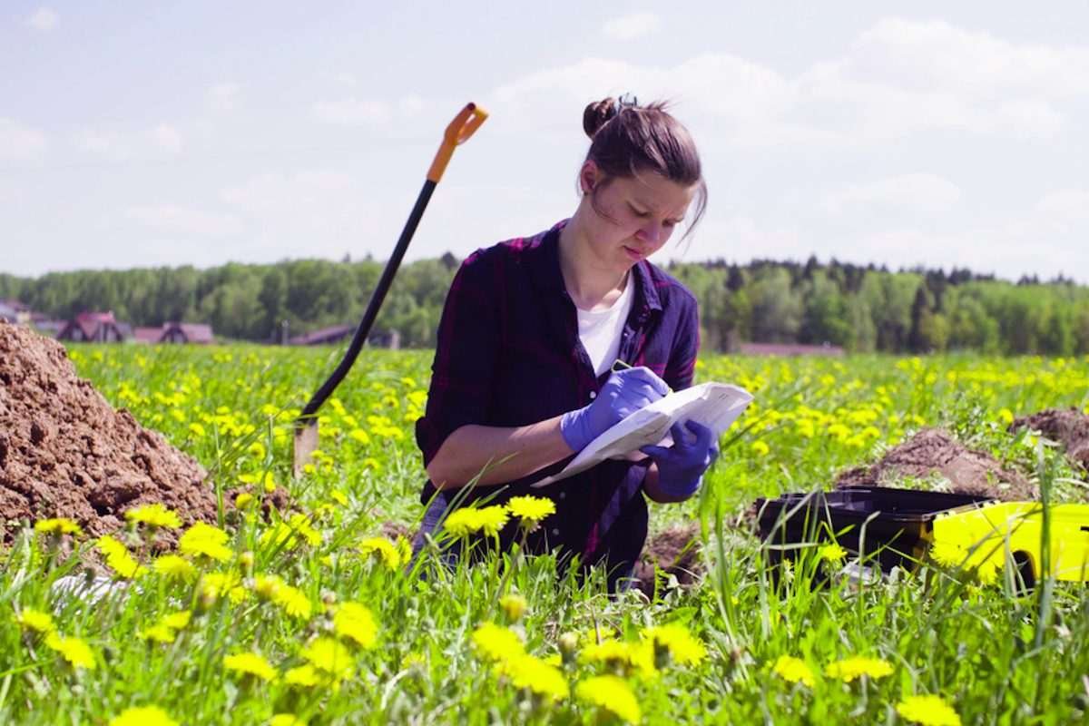 A woman stands in a field surrounded by long grass, taking notes