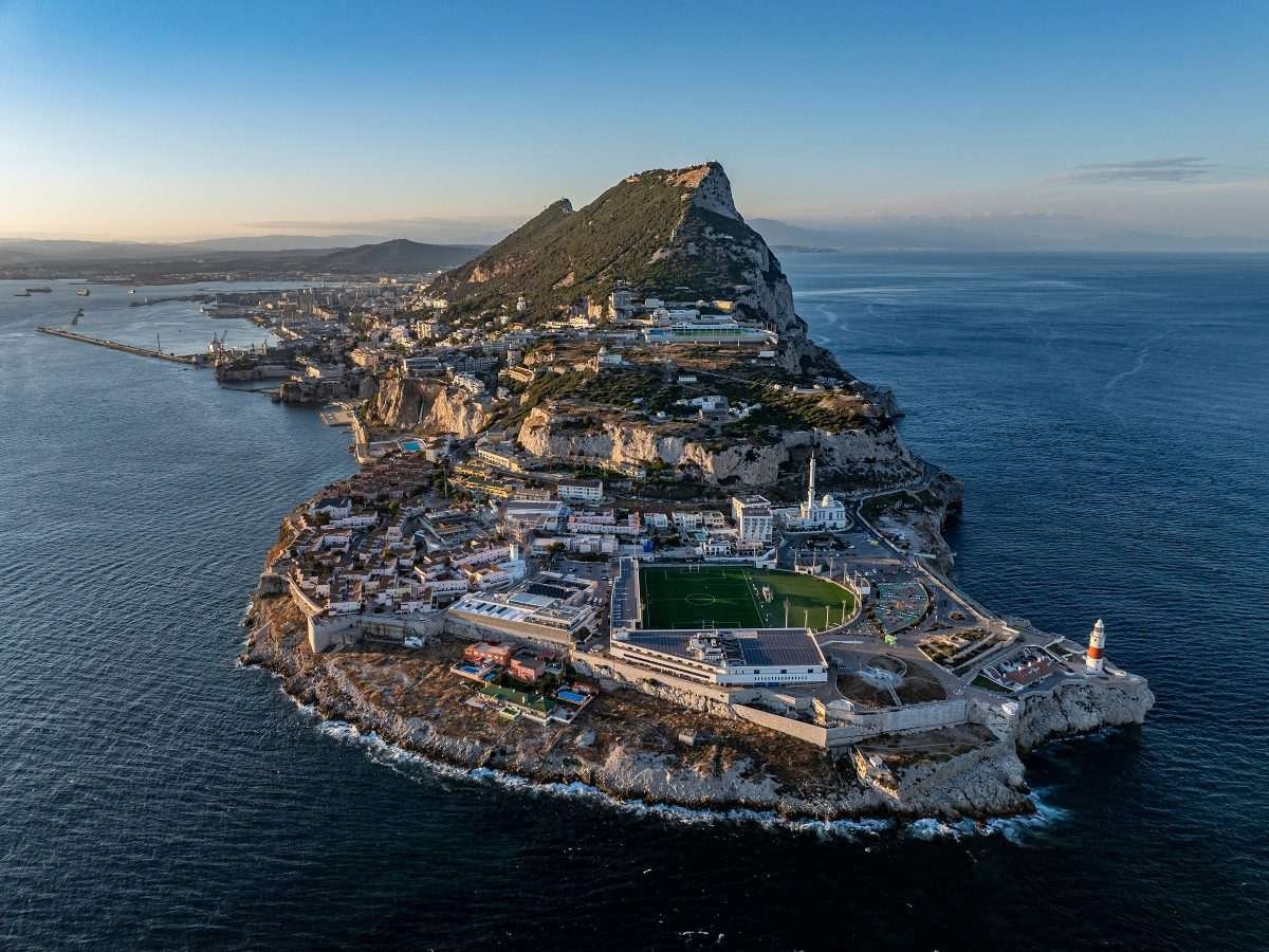 Aerial view of Gibraltar, viewed from a sea location beyond the outermost point of the peninsula, displaying sunrise view of buildings and other features arrayed below, with the rock in the distance