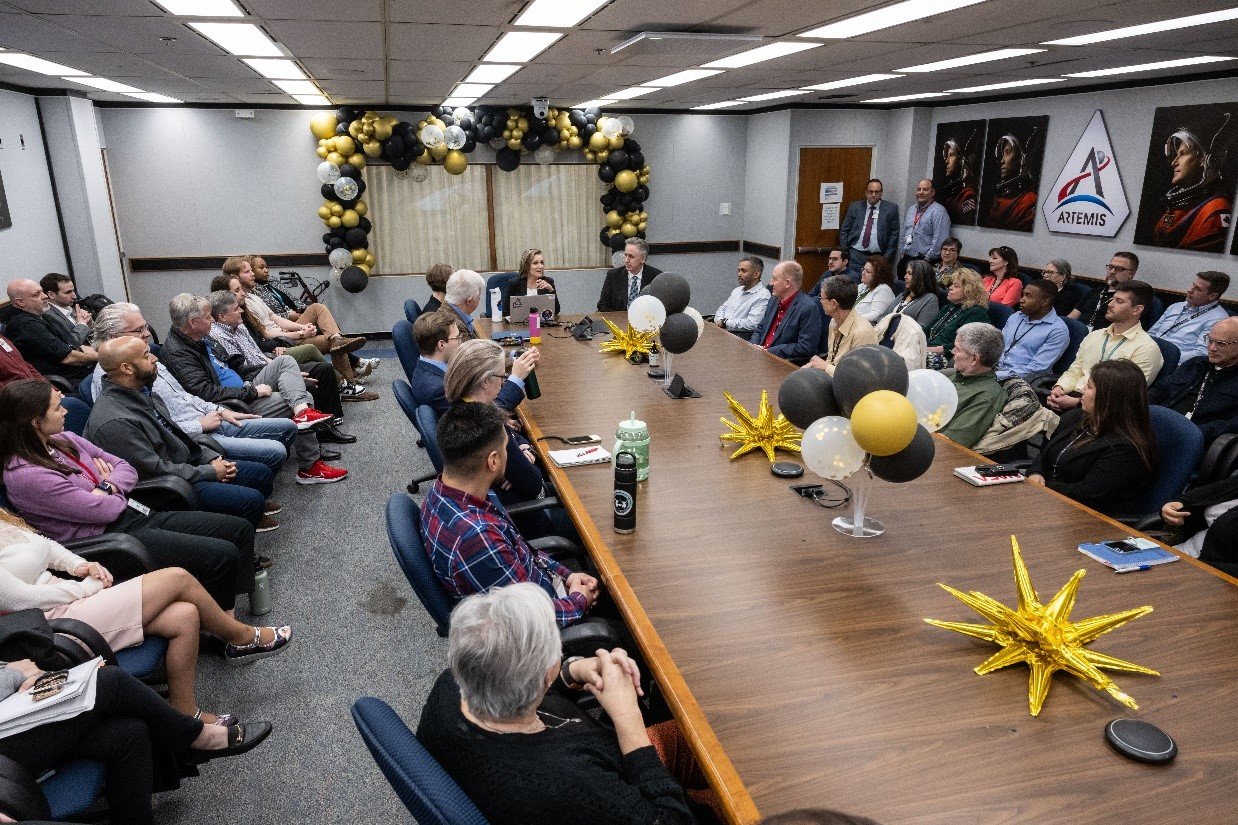 Fourteen people of different ages and races sit around a conference table, with another 20 individuals seated in chairs to either side of the table, while they participate in a team meeting.