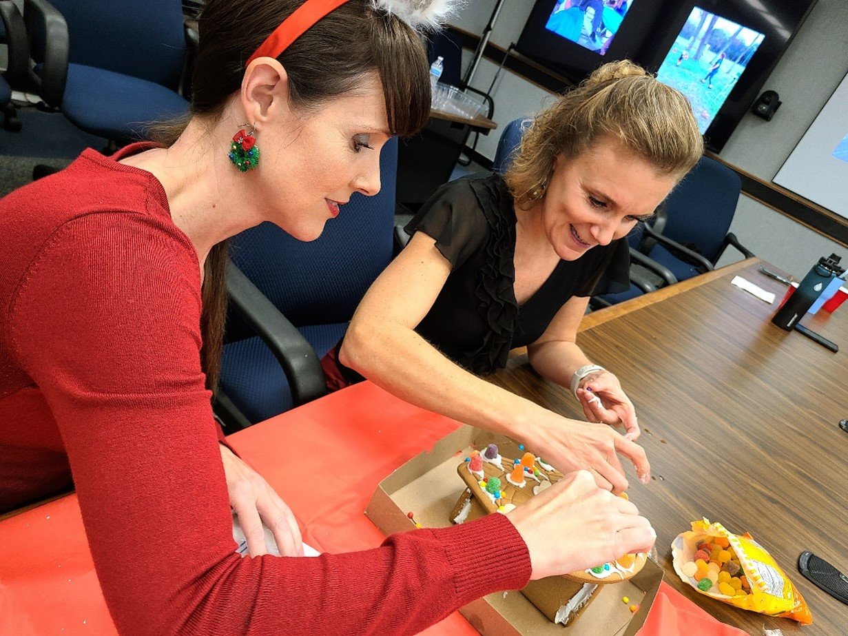 Two women in professional attire sit at a conference room table, decorating a holiday gingerbread house.