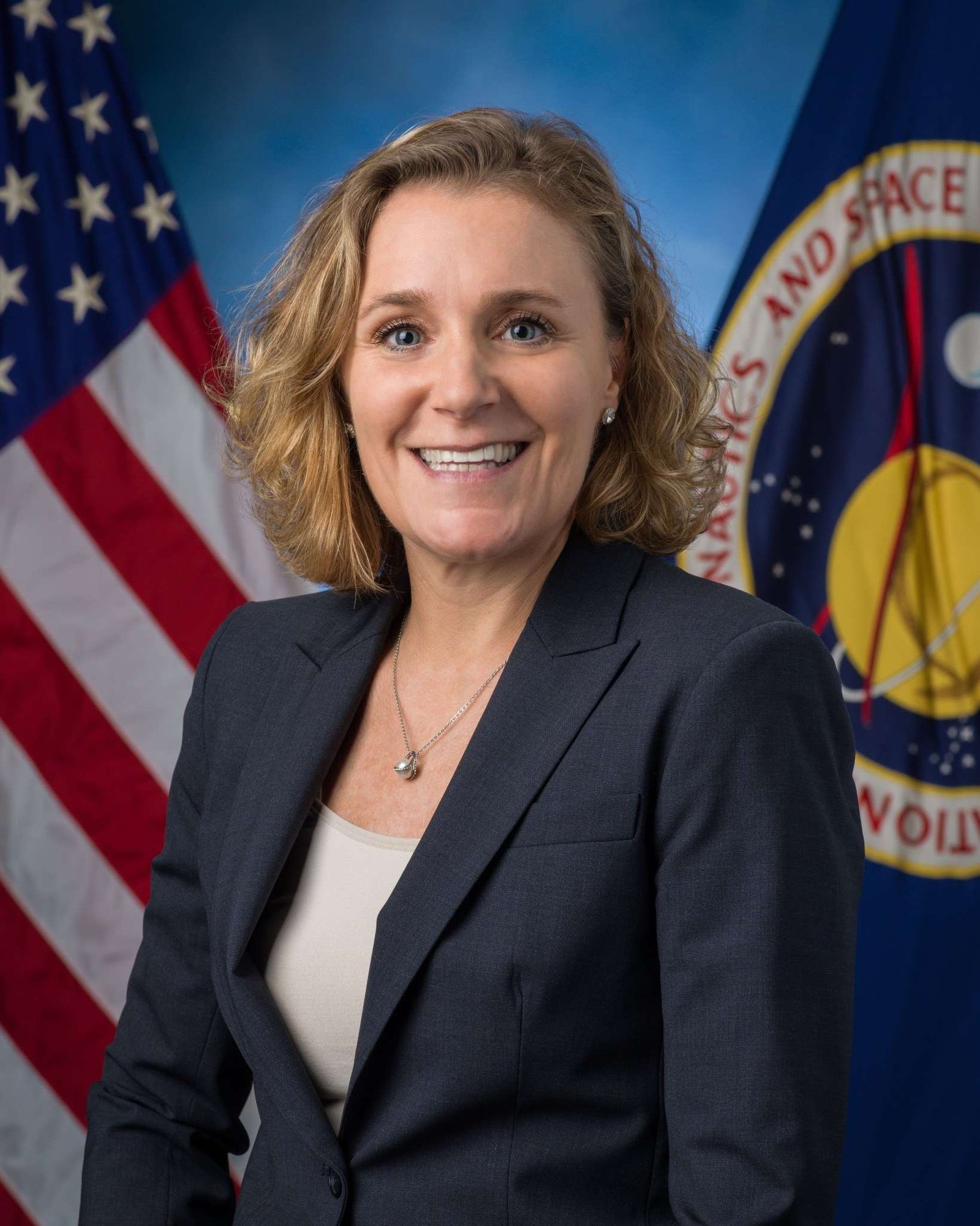 A woman with short, curly blonde hair, wearing a dark gray blazer and a beige blouse, sits in front of a NASA and an American flag.
