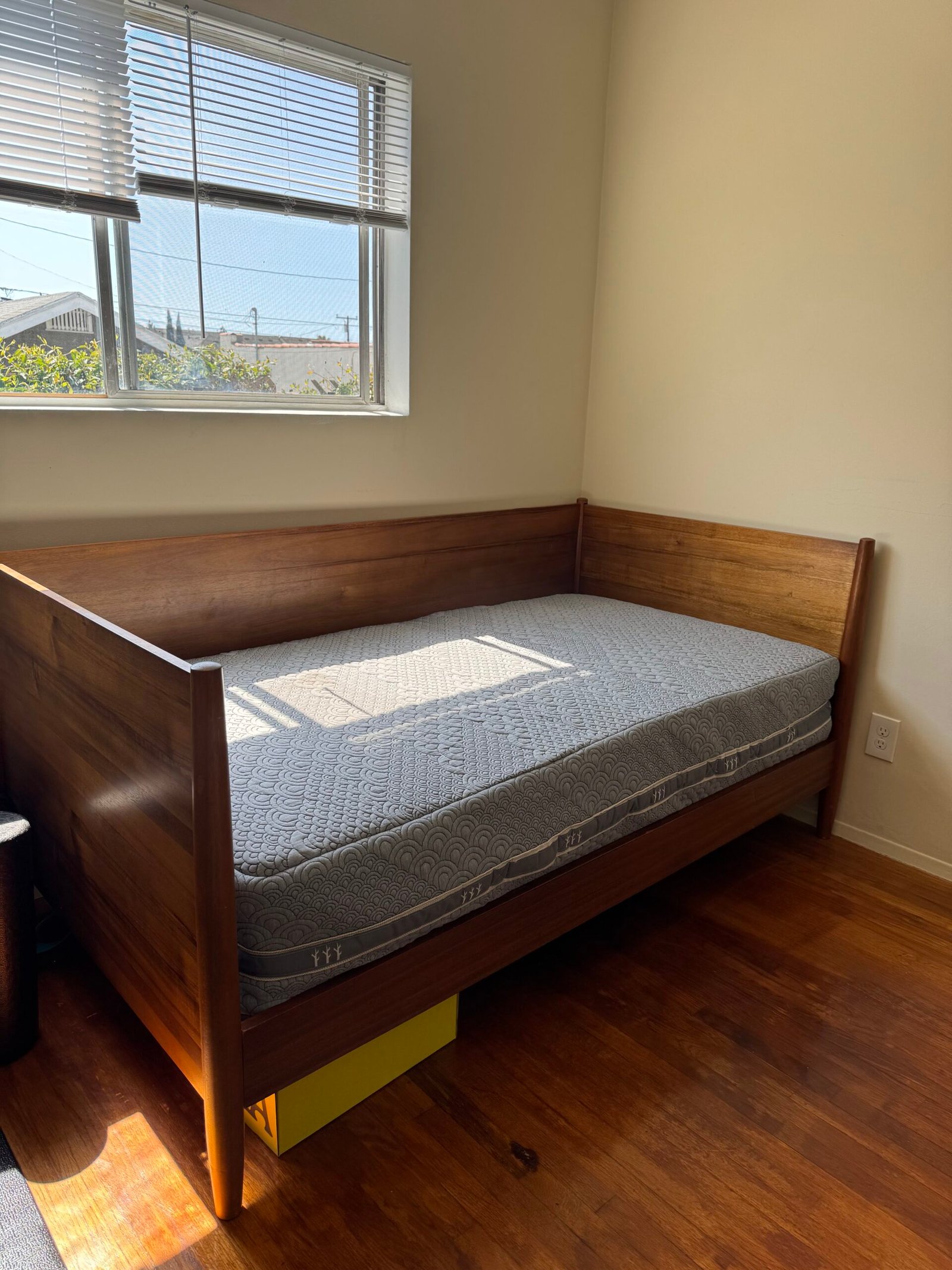 A wooden daybed with a mattress is positioned in the corner of a sunlit room with hardwood floors and a window with blinds.