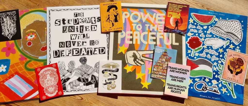 A series of artistic posters on a table, one advocating for queer rights, the next student encampments reading 'the Students united will never be defeated'. The next sign says 'Power to the Peaceful', the next advocates for a free Palestine. On top of the posters various social justice stickers are scattered.