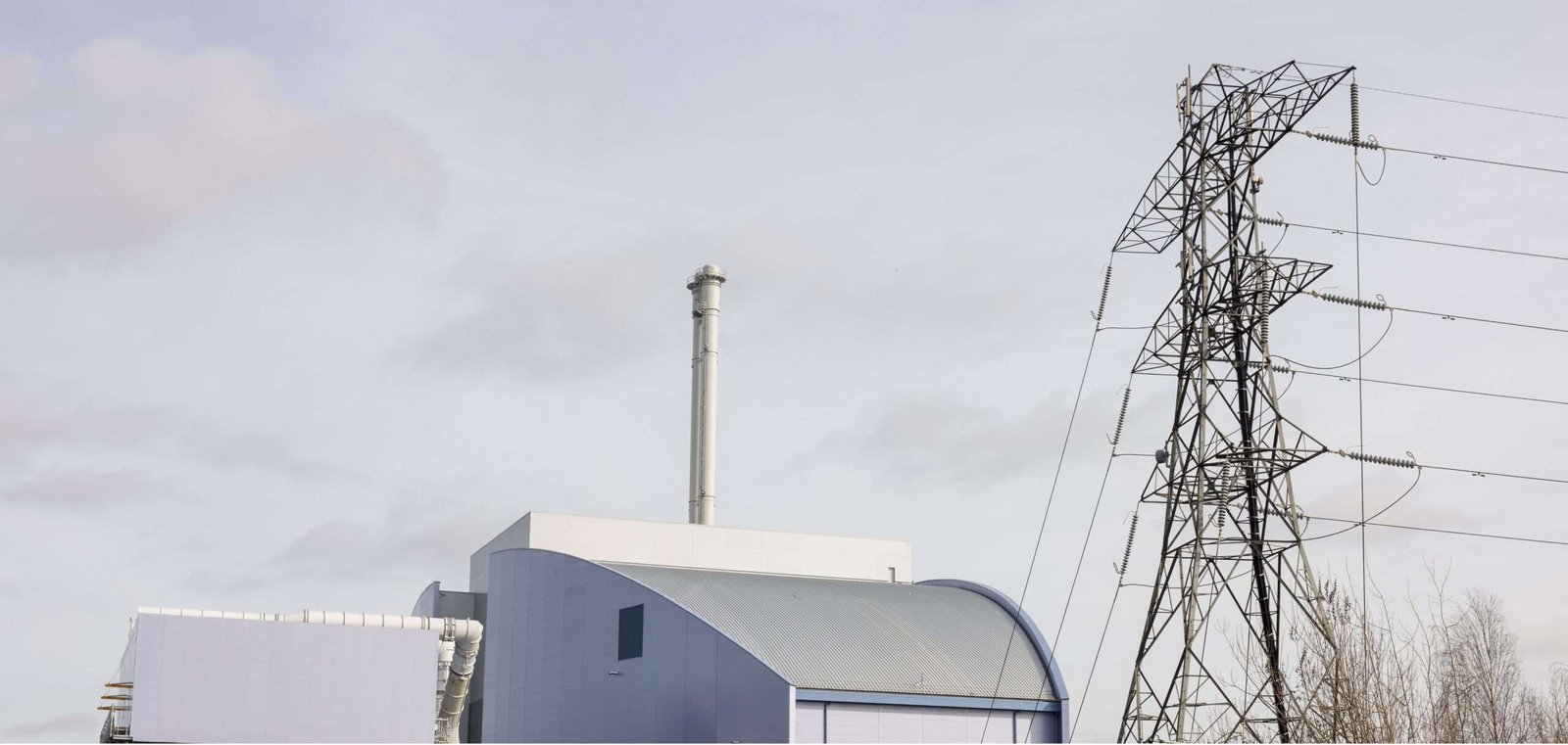 Image showing the upper portion of an energy-from-waste facility, being a cuboidal construction with a curved-slope roof and a tall cylindrical chimney, all set against a pale or grey sky background, and with an electricity transmission pylon visible to the right of the image