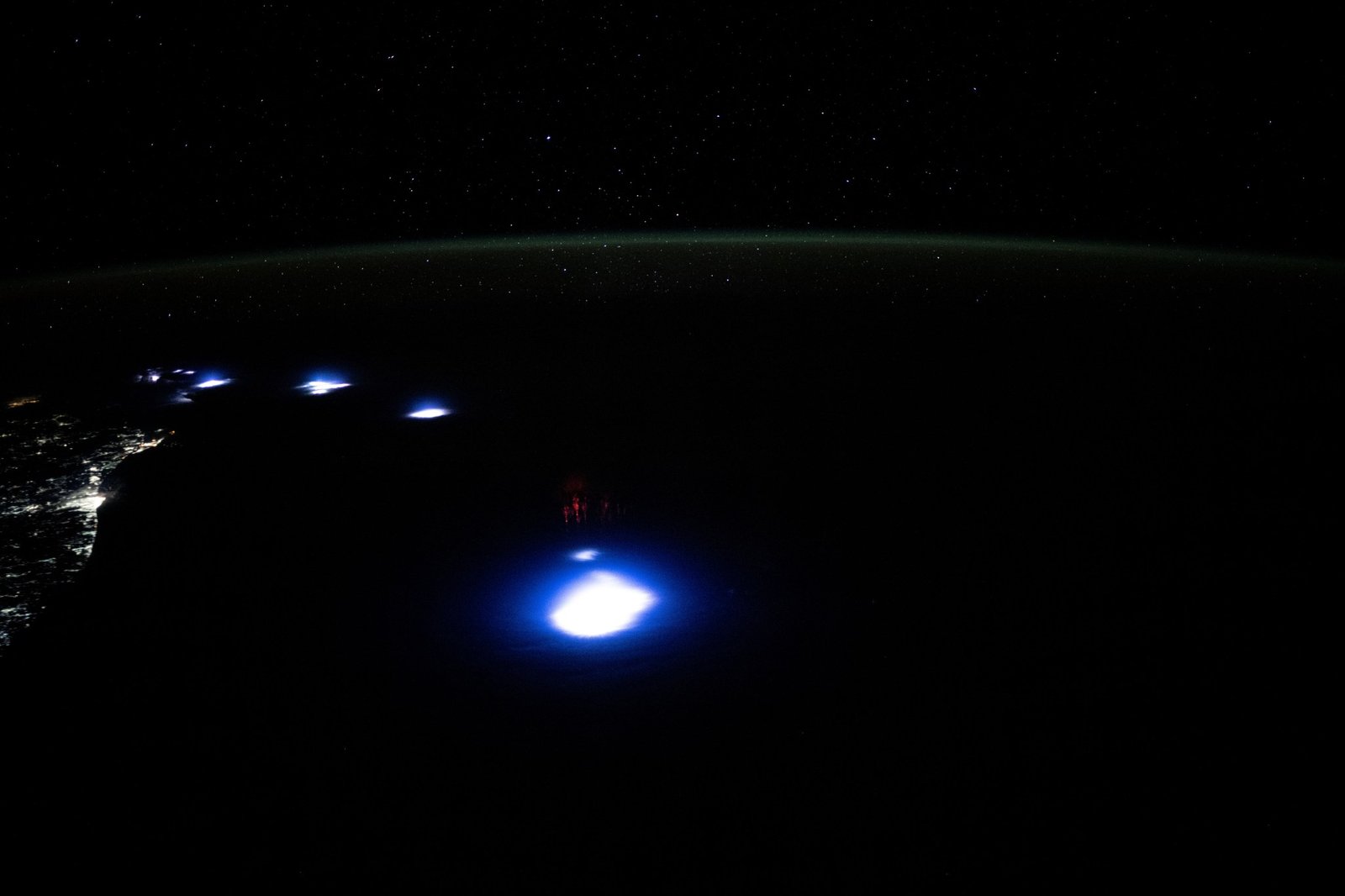 Lightning creates a large, bright white spot in the center of this image, with several sprites, thin red vertical lines, above it. Several other white spots extend to the left and the city lights line the coast on the far left. Earth’s horizon is a faint green band across the top of the image.