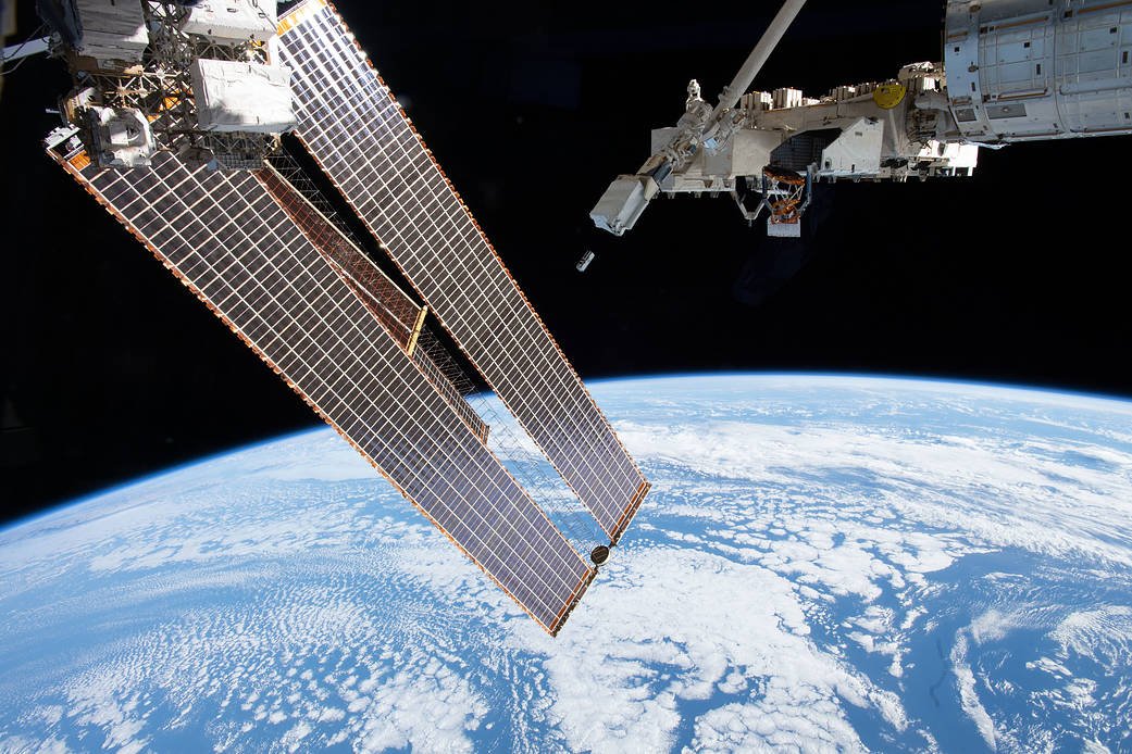A module of the space station is visible in the upper right of this image, with an arm extending downward toward the middle of it. At the end of the arm is a white square deployer with two boxes on its right side. The door on one of the boxes is open and just below it is a white and black rectangular CubeSat that has just been ejected from the deployer. Long, gold-colored solar panels fill the left side of the image and Earth covers the lower half of the image, blue ocean with scattered white clouds.