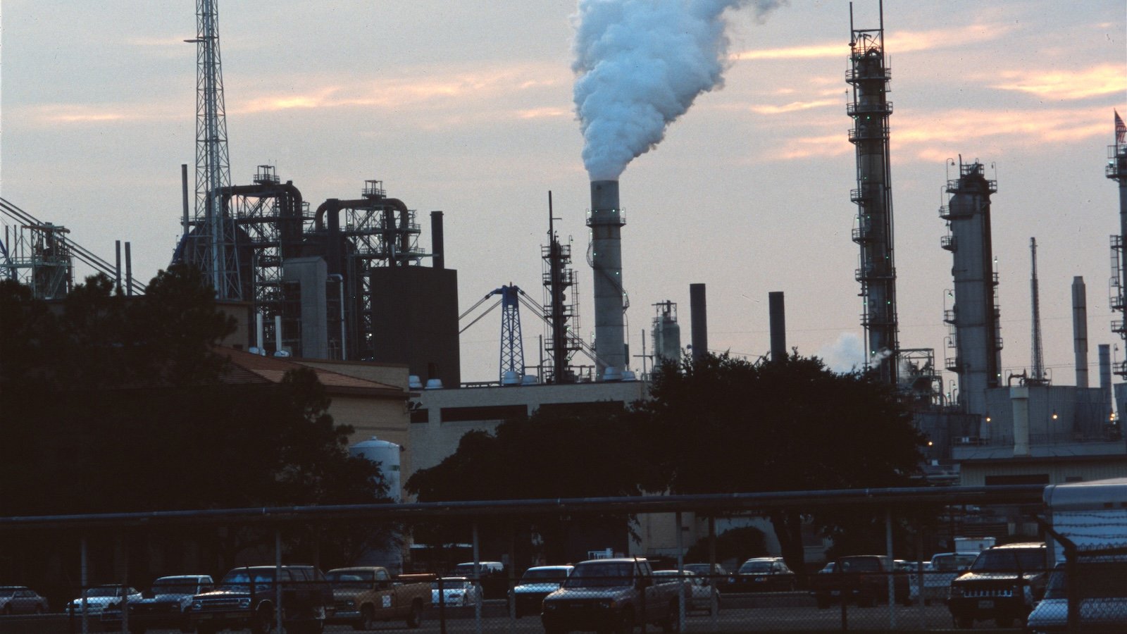 Dark view of a factory with plume emitting from a chimney and cars parked in the parking lot.